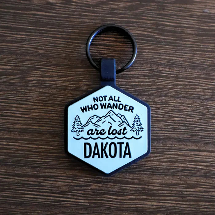 Hexagonal silicone pet ID tag with a 'Not All Who Wander Are Lost' design and the name 'Dakota' engraved on the front on a wooden surface