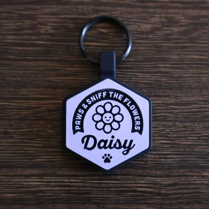 Hexagonal silicone pet ID tag with a 'Paws & Sniff the Flowers' design and the name 'Daisy' engraved on it, on a wooden surface