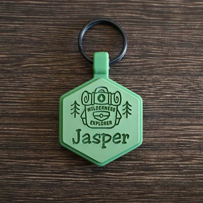 Hexagonal silicone pet ID tag with a 'Wilderness Explorer' design and the name 'Jasper' engraved on it, on a wooden surface