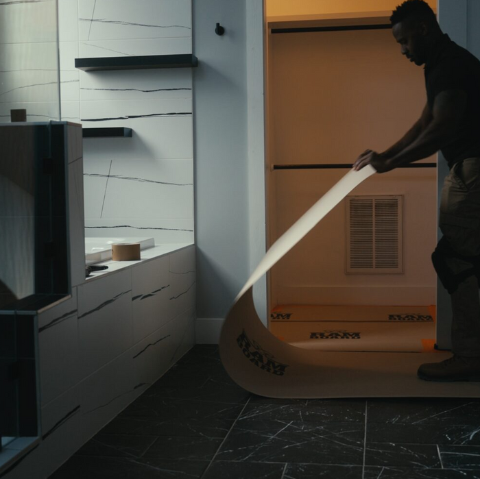 A man in a dark room is unrolling a long, curved beige adhesive strip or mat with black printed text, positioned near a modern white kitchen counter and dark tiled floor.