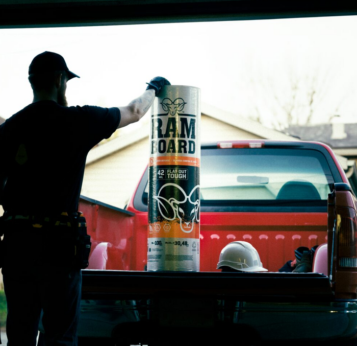 A person in a black shirt and cap stands in the bed of a red pickup truck holding a large Ram Board flat post tube. The truck bed also contains a white hard hat, with a house and bright sky in the background.