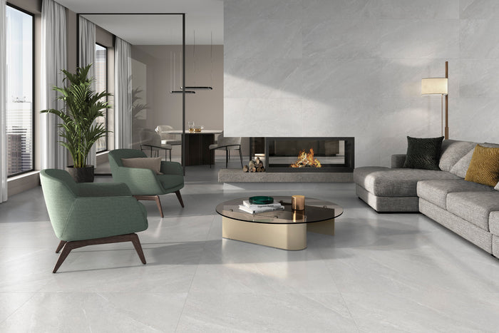 A modern living room with light gray stone-look porcelain floor tiles, featuring a gray sectional sofa, two green accent chairs, a round coffee table, and a built-in fireplace against a textured gray wall.