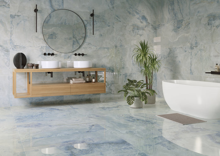 A modern bathroom interior with blue-gray marble-look tiles, a floating light wood double-sink vanity, a round wall mirror, a freestanding white bathtub, and potted green plants.