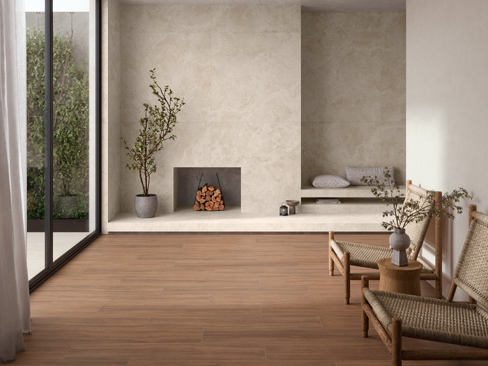 A modern interior room with warm medium-brown wood plank flooring, textured beige walls, a built-in fireplace with stacked logs, woven chairs, and potted plants.